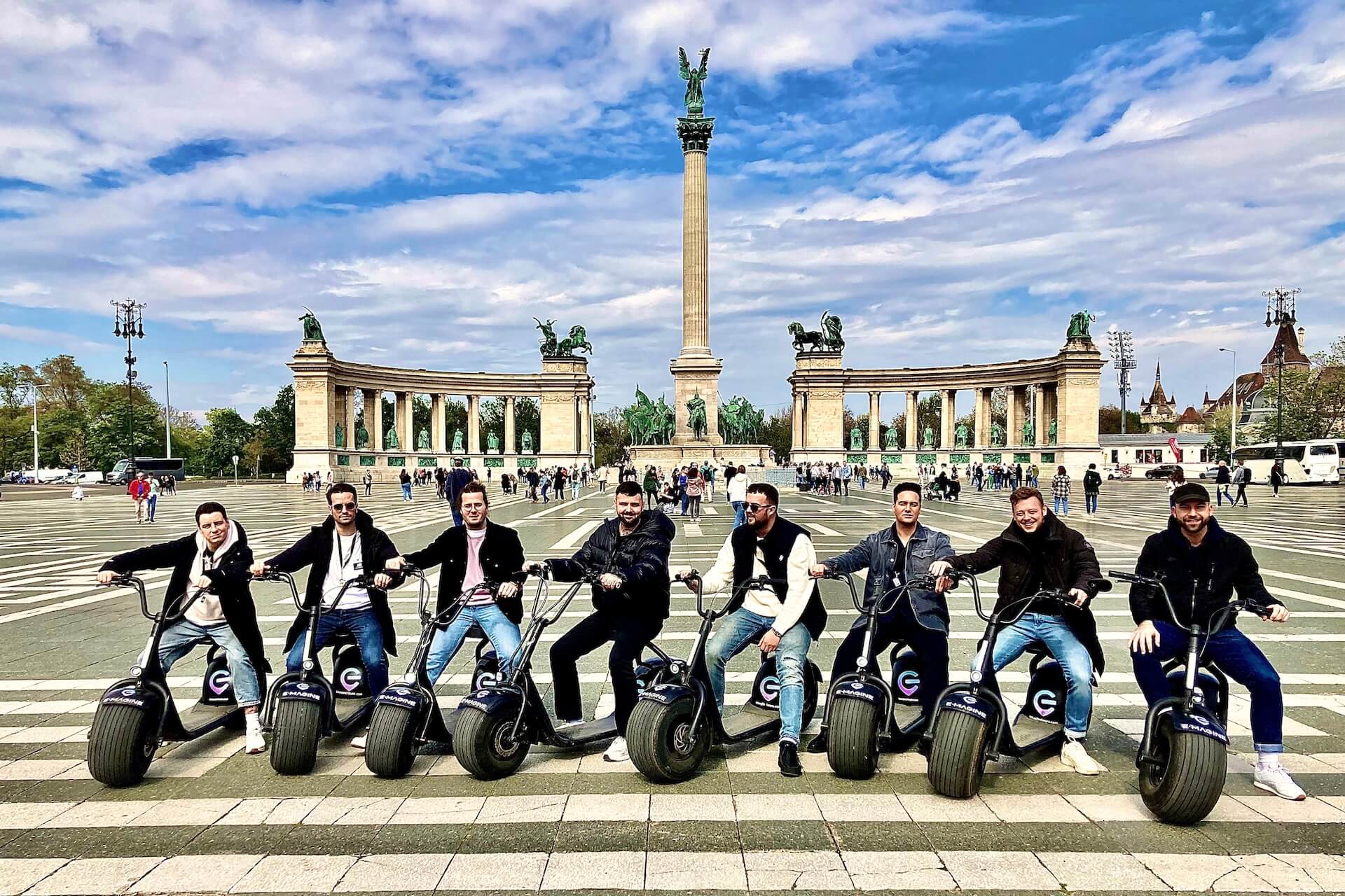 People posing on e-scooters in front of Heroes' Square monument.