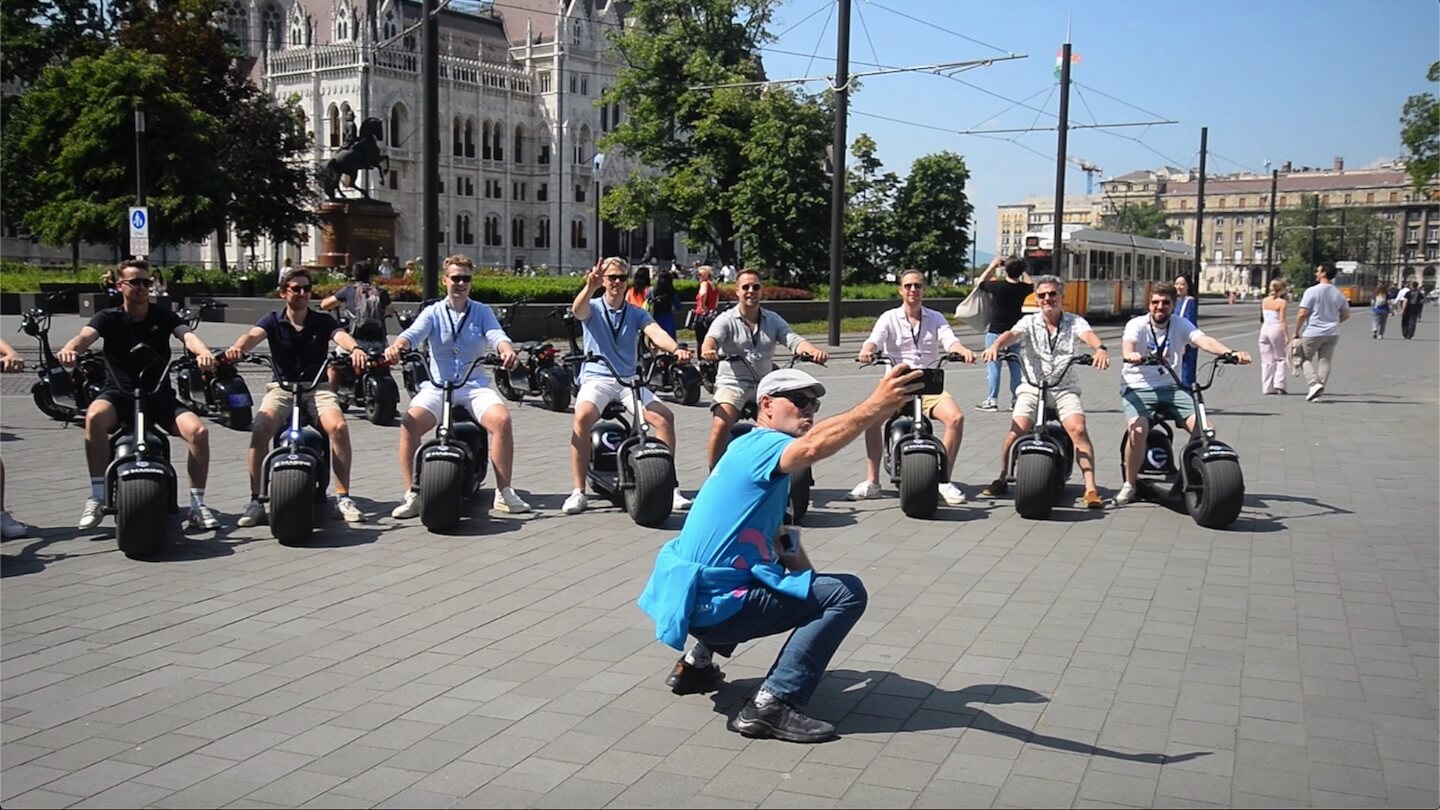 egy csapat srác e-robogón fényképezkednek a Parlament előtt Budapesten