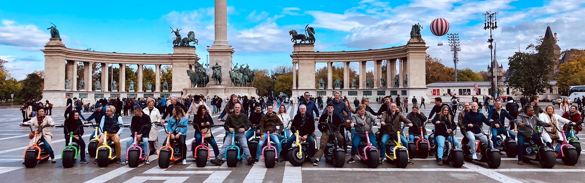 a group of riders on monsteroller e-scooters in front of the Budapest Heroes' Square monument