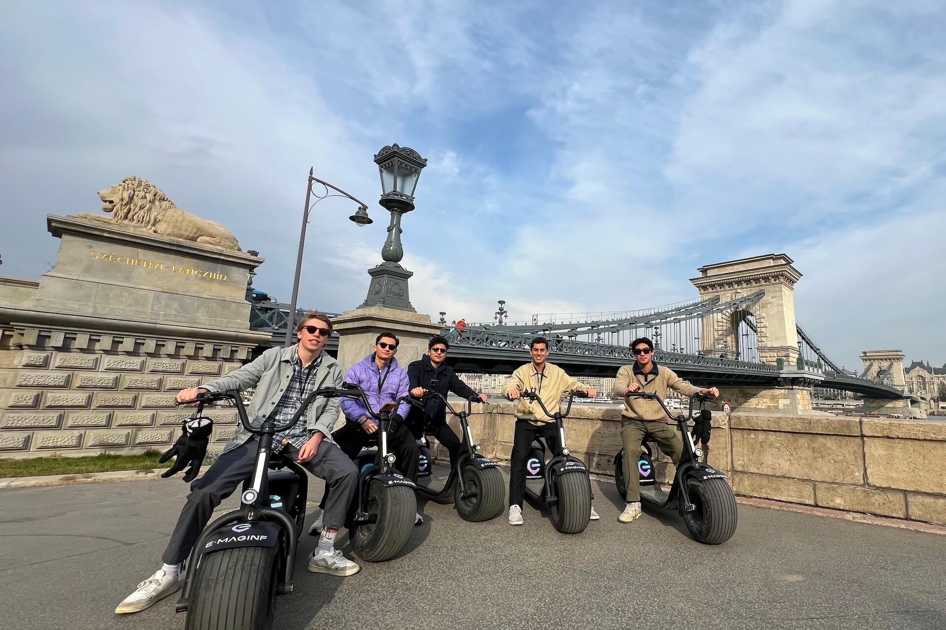 guys sitting on escooters in front of the chain bridge in budapest